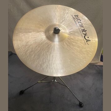 Zlatavý kovový činel umístěný na chromovaném stojanu. Na povrchu je černý nápis „Istanbul“ s hvězdicí a pod ním menší text „Mehmet Handmade Cymbals f…