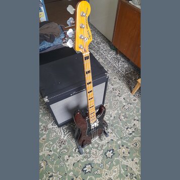 Elektrická basová kytara s tmavě hnědým trupem a hnědým pickguardem. Na hlavě nástroje je nápis Squier PRECISION BASS. Krk má světle hnědou barvu s v…
