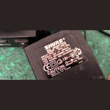 Černý síťový adaptér s bílým textem. Je na něm uveden nápis SHURE® AC ADAPTER, MODEL NO: PS31E, INPUT: 230V ~ 50Hz 0.06A a OUTPUT: 15V = 400mA. Dále…