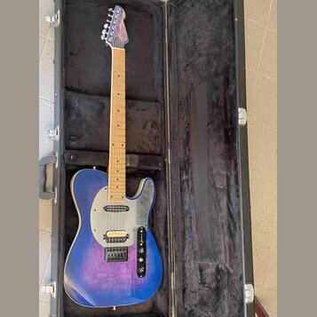 Modrá elektrická kytara typu Telecaster s javorovým hmatníkem v černém tvrdém kufru. Na korbisku je viditelné logo "Fender". Kytara má stříbrný plech…