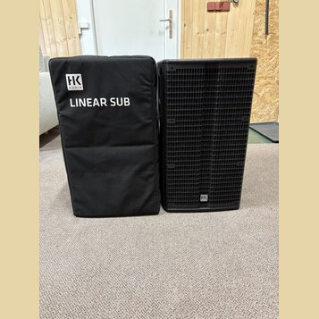 Na fotografii je černý subwoofer a přiléhající černý transportní kryt z látky. Na krytu je bílé logo „HK AUDIO“ a nápis „LINEAR SUB“. Subwoofer má če…