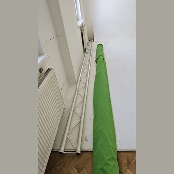 Na fotografii je stříbrný hliníkový mřížový truss v poloze vleže na podlaze. Vedle něj leží dlouhý zelený textilní kryt nebo přepravní vak. Na vybave…