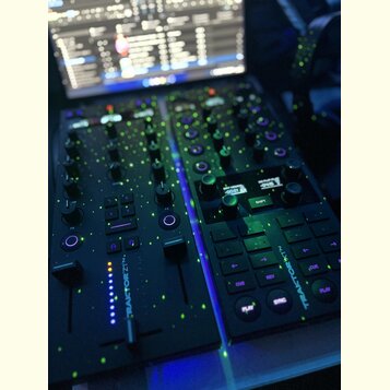 Černý DJ kontrolér s několika knoflíky, posuvníky a podsvícenými tlačítky. Na přístroji je svisle napsán bílý text TRAKTOR Z1. V pozadí je vidět zapn…