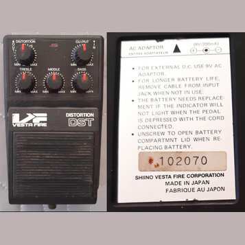 Černý efekt pedál s nápisy VESTA FIRE, DISTORTION a DST. Na horní části jsou čtyři ovládací knoflíky s popisky DISTORTION, TREBLE, MIDDLE a BASS. Dru…