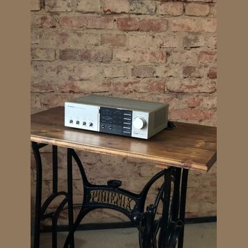 Stříbrný audio zesilovač s černým středovým panelem, umístěný na dřevěném stole. Na levé straně je nápis PIONEER a vedle něj text STEREO INTEGRATED A…