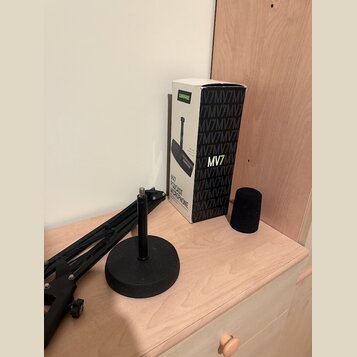 Na fotografii je krabice od mikrofonu s nápisy SHURE, MV7 a MV7 PODCAST MICROPHONE. Vedle krabice je černý pěnový popfilter a černý mikrofonní ramení…