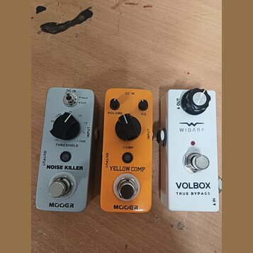 Tři efektové pedály v řadě na dřevěném povrchu. První je šedý s nápisy MOOER, NOISE KILLER a THRESHOLD. Prostřední je oranžový s nápisy MOOER, YELLOW…