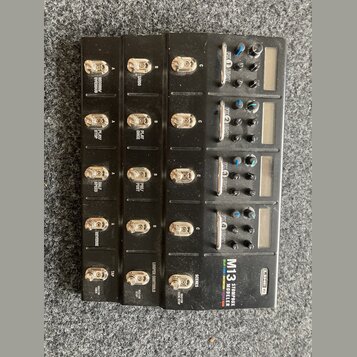 Černý multiefektový pedál s kovovým povrchem. Na spodní pravé straně je bílý nápis M13 STOMPBOX MODELER a logo LINE 6. Zařízení obsahuje pět řad spín…