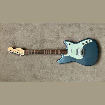 Modře šedá elektrická kytara s bílým pickguardem a hnědým hmatníkem. Na hlavě je viditelné logo "Fender". Nástroj má konfiguraci snímačů dva jednoduc…