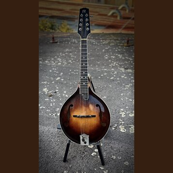 Mandolina s hruškovitým korpusem v tmavě hnědém sunburst provedení. Na hlavě je bílý nápis „12“. Nástroj stojí na černém stojanu. Povrch laku vykazuj…