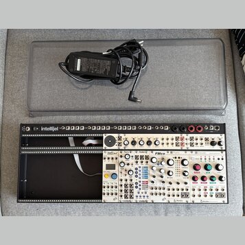 Fotografie zobrazuje Eurorack modulární syntetizátor v černém racku s bílým předním panelem a přiložený napájecí adaptér v šedém přepravním kufru. Na…