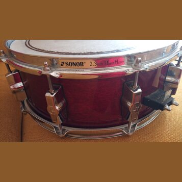 Fotografie znázorňuje snare drum s tmavě červeným dřevěným korpusem a chromovaným kováním. Na horním obruči je nápis SONOR 2.3mm PowerHoop doprovázen…