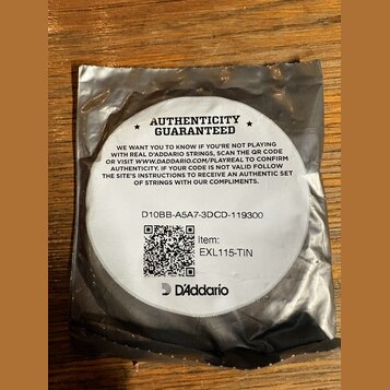 Kruhová bílá nálepka s černým textem umístěná na stříbrném obalu. Na nálepce je nápis AUTHENTICITY GUARANTEED, text s instrukcemi k ověření pravosti…