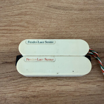Dva bílé snímače s oválným tvarem, umístěné vedle sebe na dřevěném povrchu. Oba mají nápis Fender-Lace Sensor, přičemž horní je černým písmem a dolní…