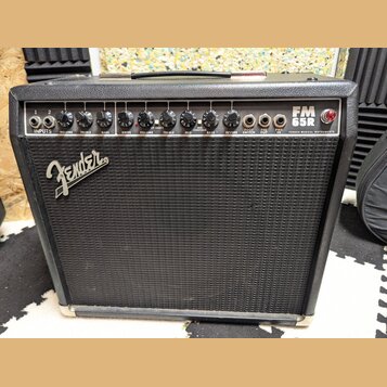 Černý kytarový kombozesilovač s textilní maskou a stříbrným logem Fender. Na horním ovládacím panelu je nápis FM 65R a FENDER MUSICAL INSTRUMENTS. Pa…
