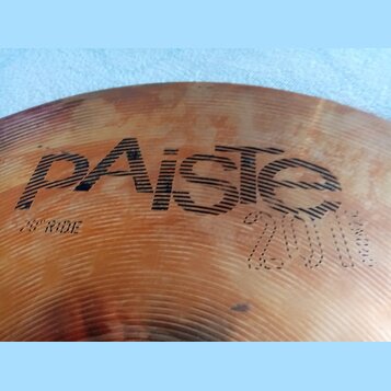 Detailní záběr na povrch činchy v bronzové barvě. Na povrchu je vidět nápis „paiste“ a pod ním označení „20" RIDE“. V pravé dolní části je vyryto „20…