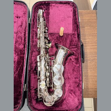 Stříbrný saxofon v černém kufru s fialovým polstrováním. Nástroj vykazuje známky opotřebení v podobě drobných škrábanců a znečištění povrchu. Na těle…
