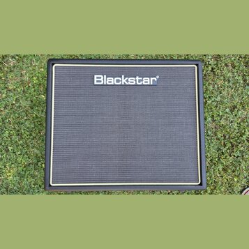 Kytarový kombozesilovač s černým povrchem a šedou textilní maskou. V horní části je umístěno stříbrné logo s nápisem Blackstar. Celý přístroj je obal…