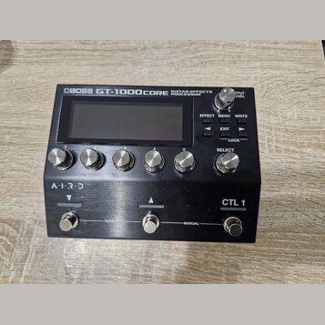 Černý kytarový procesor s nápisem BOSS GT-1000CORE GUITAR EFFECTS PROCESSOR. Zařízení má digitální displej, šest kovových otočných ovladačů s čísly 1…
