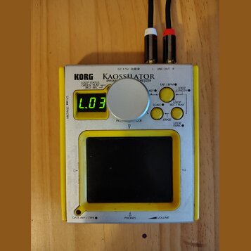 Stříbro-žlutý přenosný syntetizátor s nápisy KORG a KAOSSILATOR DYNAMIC SYNTHESIZER. Zařízení má malý digitální displej s nápisem L.03, jeden velký s…