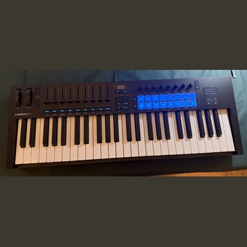 Černý MIDI kontrolér s klaviaturní klávesnicí. Na levé straně je bílý nápis LAUNCHKEY a řada posuvníků. V pravé horní části se nachází mřížka 16 modř…