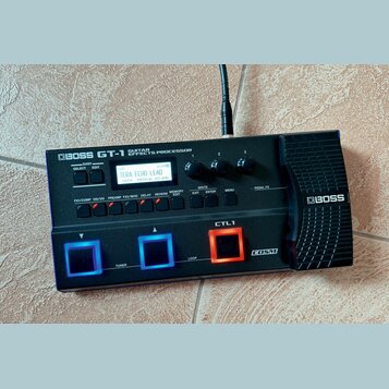 Černý kytarový procesor s nápisem BOSS GT-1 GUITAR EFFECTS PROCESSOR. Zařízení má digitální displej, na kterém je text TERA ECHO LEAD a další ovládac…