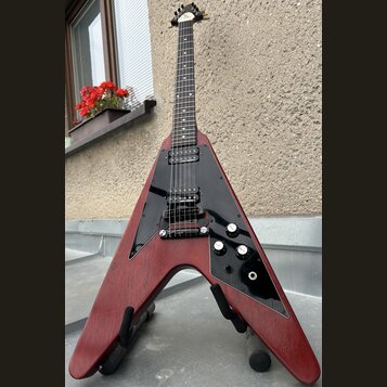 Elektrická kytara s Flying V tvarem těla v červeném dřevěném provedení a černým pickguardem. Na hmatníku jsou dva humbuckery, tři ovládací knofíky a…