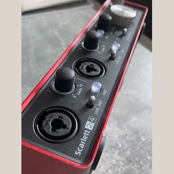 Červený audio rozhraní s černým horním panelem. Na panelu je nápis Scarlett 2i4, dva vstupy typu combo jack/XLR a ovládací prvky s popisky GAIN 1, GA…