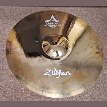 Kruhový mosazný činel s viditelným broušením povrchu. Na horní části je bílý nápis CUSTOM 20th Anniversary Ride MEDIUM THIN 21"/53 cm s doprovodným l…