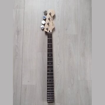 Fotografie zachycuje hmatník a hlavu basové kytary z přírodního dřeva s tmavým hmatníkem. Na hlavě je vidět nápis Squier JAZZ BASS a čtyři kovové lad…