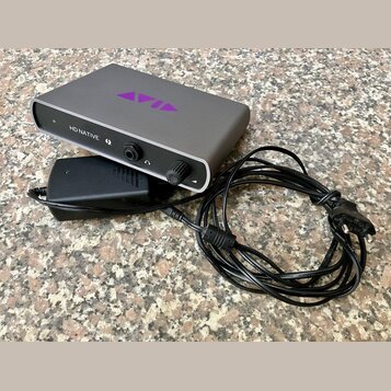 Šedý audio interface s nápisem HD NATIVE a fialovým logem AVID na horní straně. Zařízení má na předním panelu tlačítko, ovrtací knoflík a svítící kon…