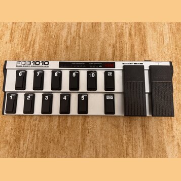 Stříbrný MIDI pedál s černými spínači a dvěma expresními pedály. Na levé straně je nápis FCB1010 MIDI FOOT CONTROLLER a nad spínači jsou sekce PRESET…