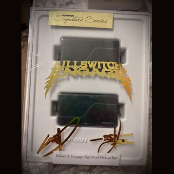 Dva černé humbuckery v bílém plastovém obalu, který je zabalen v průhledné fólii. Na horním okrape obalu je žlutý štítek s nápisem FISHMAN Signature…