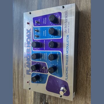 Kovový efekt pedál s nápisy FLANGER HOAX a electro-harmonix MADE IN USA. Zařízení má modré a fialové ovládací panely s několika černými otočnými knof…