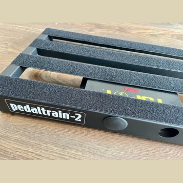 Černý kovový pedalboard s nápisem pedaltrain-2 v bílém rámečku. Horní lišty jsou potaženy šedým protiskluzovým materiálem. Pod deskou je vidět část č…