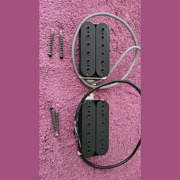 Dvě černé humbucker snímače s kovovými základnami a připojenými kabely v šedé a černé barvě. Vedle snímačů jsou položeny čtyři montážní pružiny s šro…