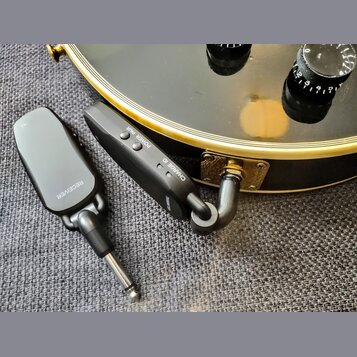 Dvě černé bezdrátové vysílače/přijímače s kovovými jack konektory. Na levém zařízení je nápis RECEIVER, na pravém jsou nápisy POWER PAIR, CHARGE ID a…