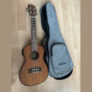 Hnědý dřevěný ukulele s doprovodným šedým textilem v podobě obalu. Na hlavě nástroje i na přední straně obalu je bílý nápis CASCHA. Na hmatníku jsou…
