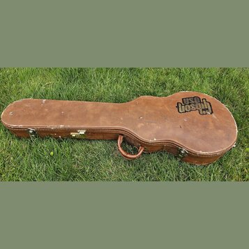 Hnědý tvrdý kufýr na kytaru s kovými zapínacími zámky a úchytem. Na pravé straně je vidět černé logo s nápisy USA a Gibson. Povrch vykazuje známky op…