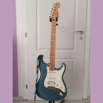 Elektrická kytara s modrým kovovým povrchem těla a světle hnědým javorovým hmatníkem. Na hlavě je viditelné logo Fender. Kytara má bílý pickguard, tř…
