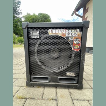 Černý basový kombozesilovač s kovovou ochrannou mřížkou přes reproduktor. Na přední straně jsou viditelná loga a nápisy: "Roland", "CUBE-120XL BASS",…