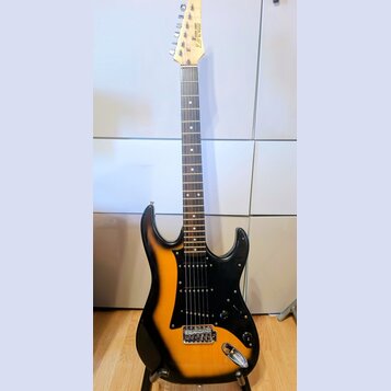 Elektrická kytara s trupem v barevně sunburst (černá a žlutá) a černým pickguardem. Na hlavě hmatníku je viditelný nápis „G&L“ a pod ním text „by Leo…