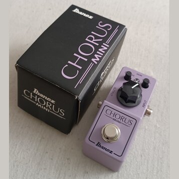 Fialový efekt pedál s nápisy CHORUS MINI a Ibanez. Na horní části je jeden černý ovládací knoflík a konektory OUTPUT a INPUT. Vedle pedálu je černá k…