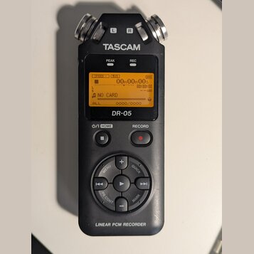 Černý digitální záznamový zařízení s nápisy TASCAM a DR-05. Displej zobrazuje hlášku NO CARD a časovač 00H00M00S. V dolní části je nápis LINEAR PCM R…