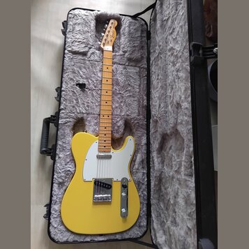 Žlutá elektrická kytara typu Telecaster s bílým pickguardem a javorovým hmatníkem v černém polstrovaném kufru. Na hlavě nástroje je viditelné logo Fe…