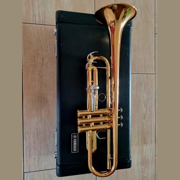 Zlatý mosazný trumpet položený na černém kufru. Na kufru je bílý nápis YAMAHA v obdélníkovém rámečku. Nástroj vykazuje známky používání v podobě drob…