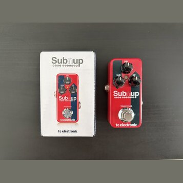 Fotografie zobrazuje červený efekt pedál a jeho originální krabici. Na pedálu i na krabici je bílý nápis Subnup MINI OCTAVER a logo tc electronic. Pe…