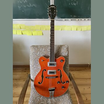 Oranžová polodupatá elektrická kytara s hnědým hmatníkem a bílým lemováním. Na hlavě je bílý nápis GRETSCH a na spodní části korpusu je kovový most s…