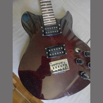 Elektrická kytara s tmavě červeným metalizovaným povrchem a dvěma černými humbuckery. Na kovovém mostku je vyryt nápis ALIEN WORKSHOP. Nástroj má tři…