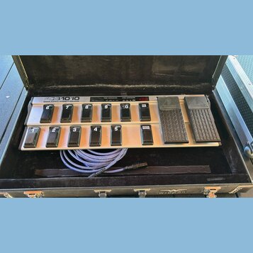Stříbrný MIDI pedál s černými spínači a dvěma expresními pedály, umístěný v černém kufru. Na horní části je nápis FCB1010 MIDI FOOT CONTROLLER a digi…